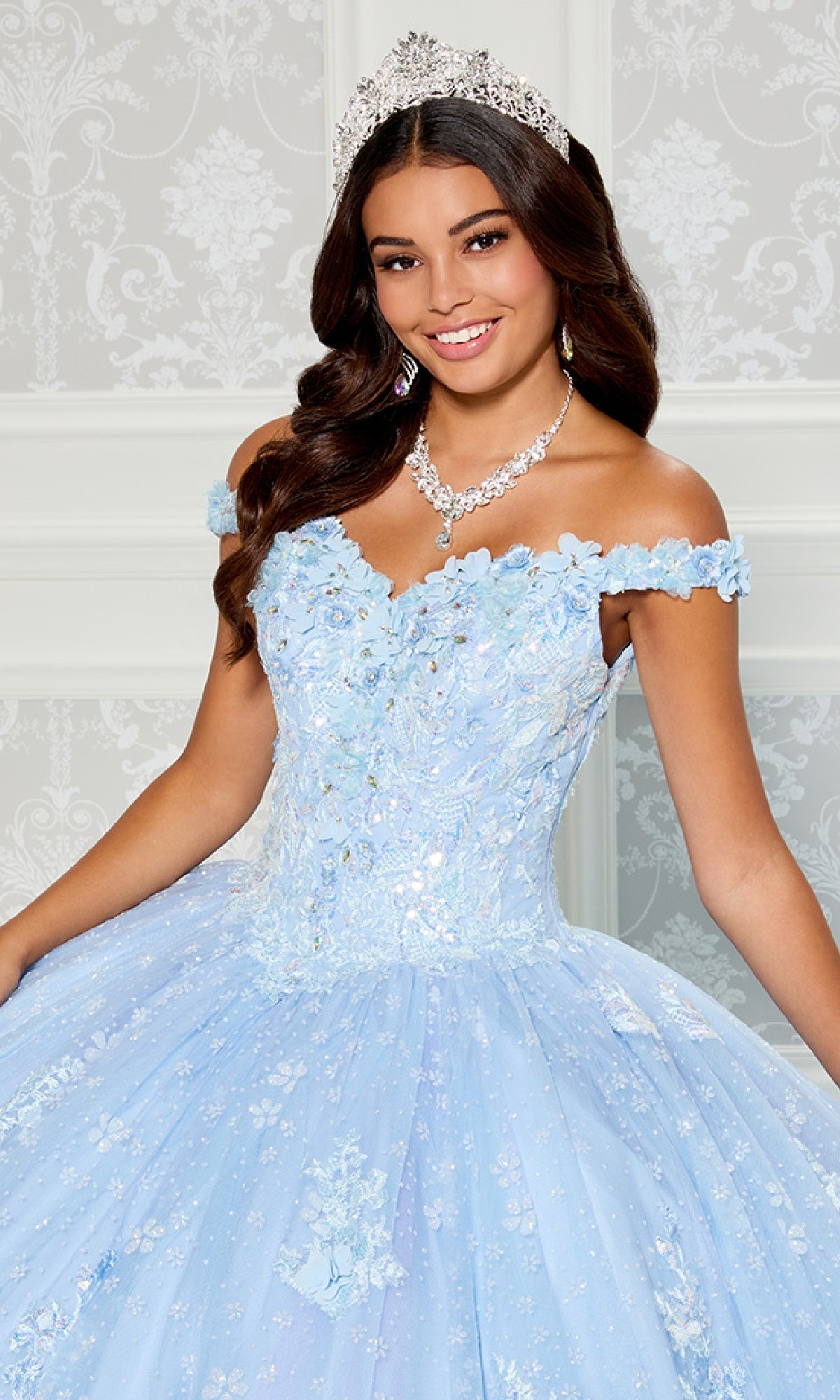 A young woman wears the Quinceanera Dress PR30111 By Princesa, a light blue off-the-shoulder gown with floral details and lace embroidery, accessorized with a jeweled tiara and matching necklace, standing by a patterned wall.